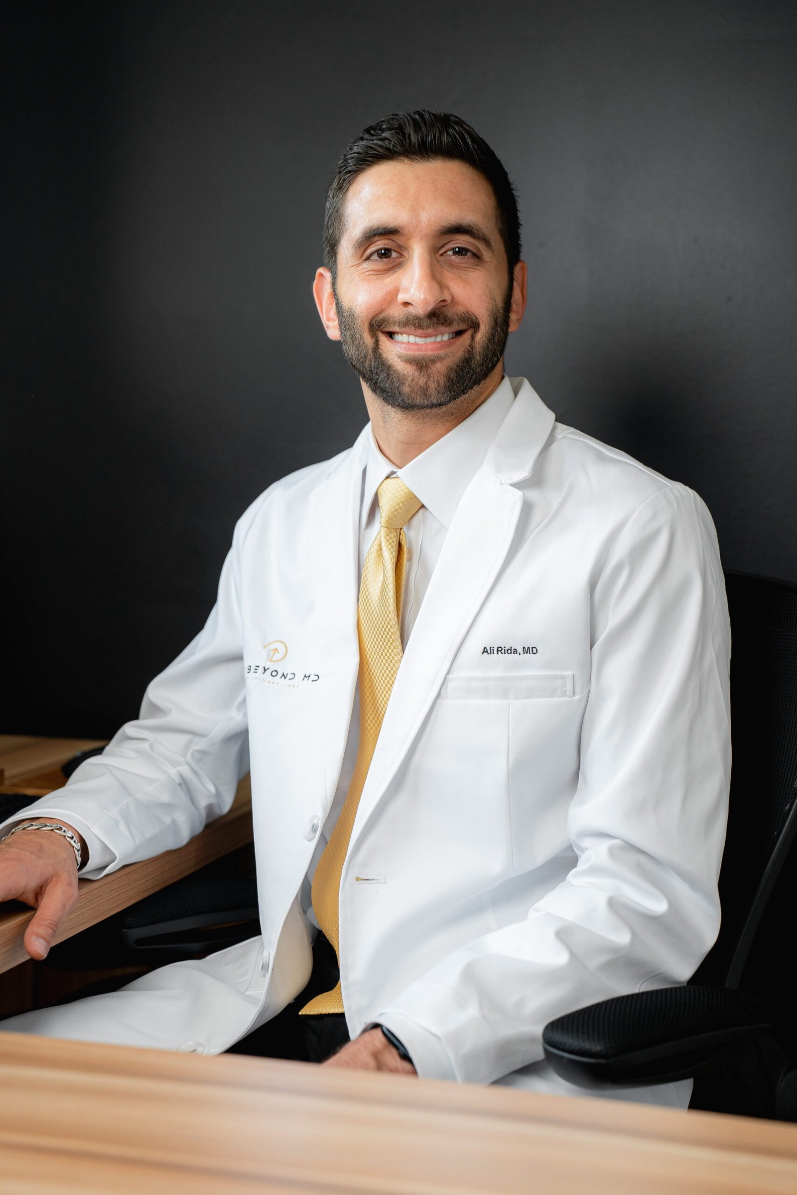 Dr. Ali Rida, board-certified family medicine physician, smiling in a white coat, seated at a desk, representing personalized primary care at Life Beyond MD Primary Care in Dearborn, Michigan.