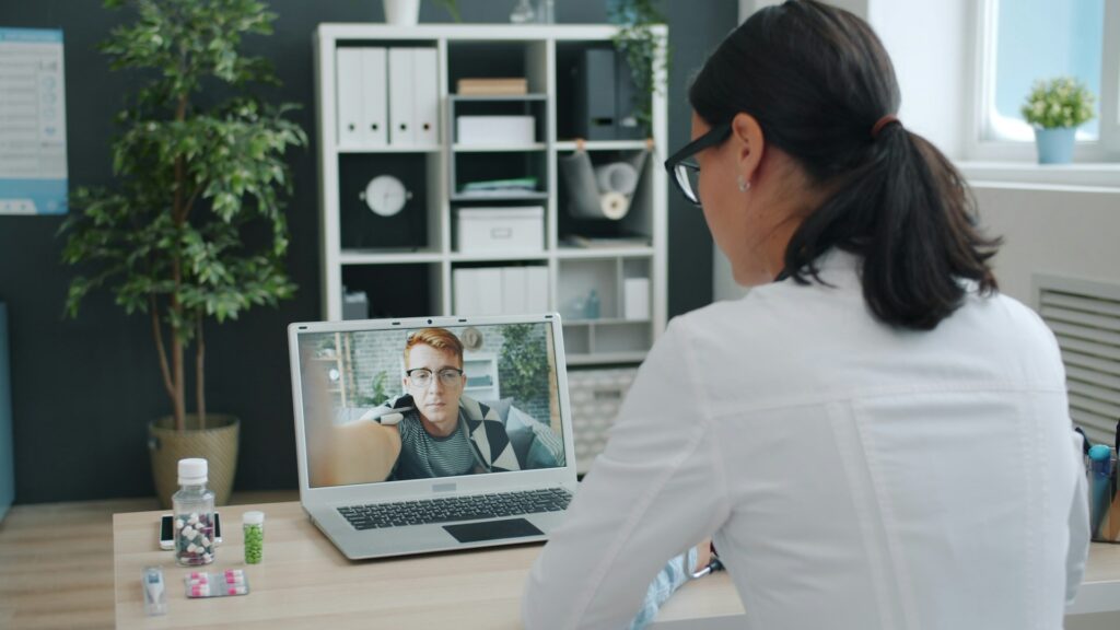 Doctor consulting with a patient via telehealth on a laptop, showcasing virtual primary care services, with a plant and medication visible in the background.